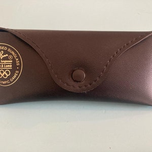 Op de afbeelding: Een bruin leren Ray-Ban zonnebril etui met gouden letters die "Officially Licensed Sunglasses Ray-Ban Sunglasses Bausch & Lomb 1984 Olympic Games" lezen.