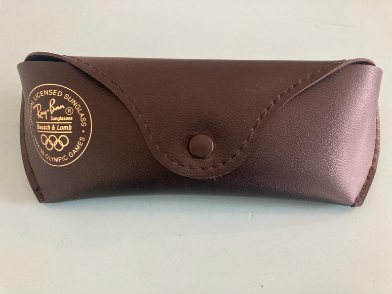 Op de afbeelding: Een bruin leren Ray-Ban zonnebril etui met gouden letters die "Officially Licensed Sunglasses Ray-Ban Sunglasses Bausch & Lomb 1984 Olympic Games" lezen.