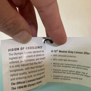 Op de afbeelding: Een wit label met zwarte tekst dat de Vision of Excellence voor de Olympische Spelen van 1994/96 beschrijft. Het label beschrijft ook de G-15 Neutral Gray Lenses en hun eigenschappen, waaronder 100% UV-bescherming, 85% absorptie van zichtbaar licht, neutrale kleurtransmissie, precisiegeslepen optisch glas en geschiktheid voor algemeen gebruik en buitenshuis.