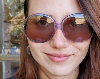 May include: A woman with freckles wearing large, round, purple-tinted sunglasses with a nose ring.