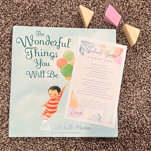 May include: A pink and blue card with the text "The School Year is Almost Done!"  The card has a watercolor background and features a message about the end of the school year. The card is sitting on top of a book titled "The Wonderful Things You Will Be" by Emily Winfield Martin.