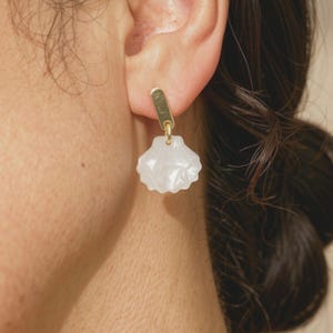 May include: A gold-toned metal earring featuring a small, vertical rectangular post attached to a translucent, white, shell-shaped pendant. The pendant has a ribbed, scalloped texture and hangs from a small circular jump ring connected to the post.