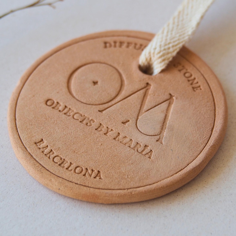May include: A circular, light brown diffuser stone with embossed text reading "DIFFUSER STONE" and "OBJECTS BY MARIA BARCELONA". It has a cream ribbon for hanging.