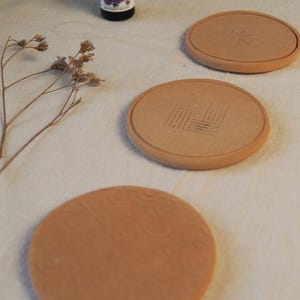 May include: Four round, terracotta-coloured coasters with etched designs. One coaster has a geometric pattern, while others feature a floral design. The coasters are arranged on a light-coloured, textured surface.