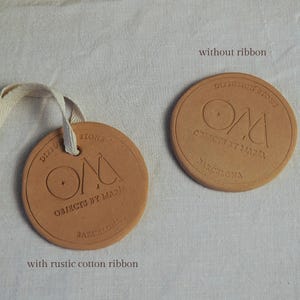 May include: Two circular terracotta-coloured objects with embossed text. One has a rustic cotton ribbon attached. The text reads "Diffusion Stone, Objects by Maria, Barcelona." The other is without a ribbon.