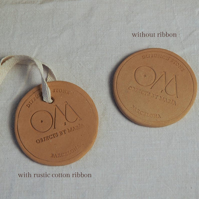 May include: Two circular terracotta-coloured objects with embossed text. One has a rustic cotton ribbon attached. The text reads "Diffusion Stone, Objects by Maria, Barcelona." The other is without a ribbon.
