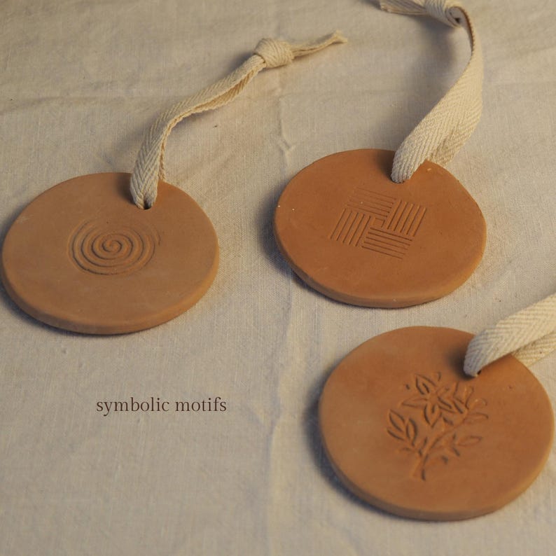 May include: Three circular terracotta ornaments with embossed designs and cream fabric loops. One has a spiral, another a geometric pattern, and the third a floral motif. The words "symbolic motifs" are printed below the ornaments.