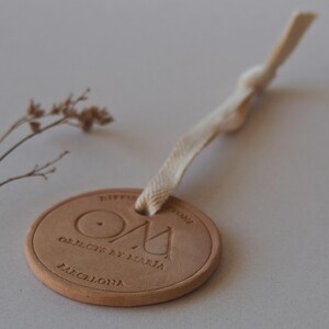 May include: A circular, tan-coloured tag with embossed text and a cream-coloured ribbon. The tag reads "OBJECTS BY MARIA" and "BARCELONA". The ribbon is knotted at the top. A sprig of dried flowers is in the background.