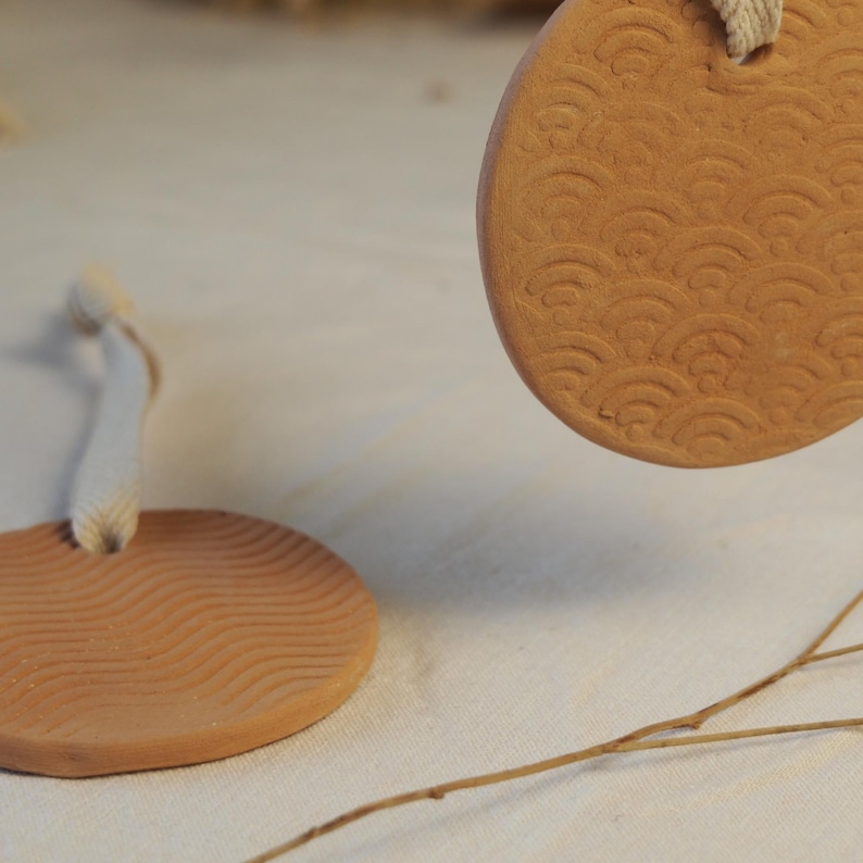 May include: Two terracotta-coloured coasters with a wave pattern are suspended against a neutral background. One coaster is in the foreground, while the other is partially visible in the background. Each coaster has a white rope attached.