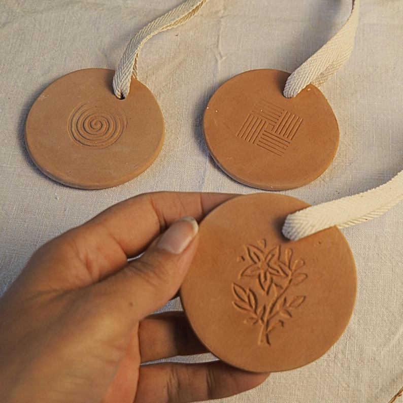 May include: Three circular terracotta ornaments with cream ribbon loops. Each has a different embossed design: a spiral, a woven pattern, and a floral motif. One ornament is held in a hand. The ornaments are made of terracotta.