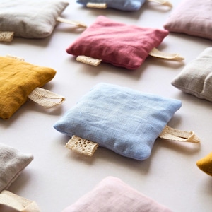 May include: A selection of small, square linen pillows in shades of yellow, pink, blue, and grey. Each pillow features a cream-coloured lace tag and a matching fabric loop. The pillows are arranged on a white surface.
