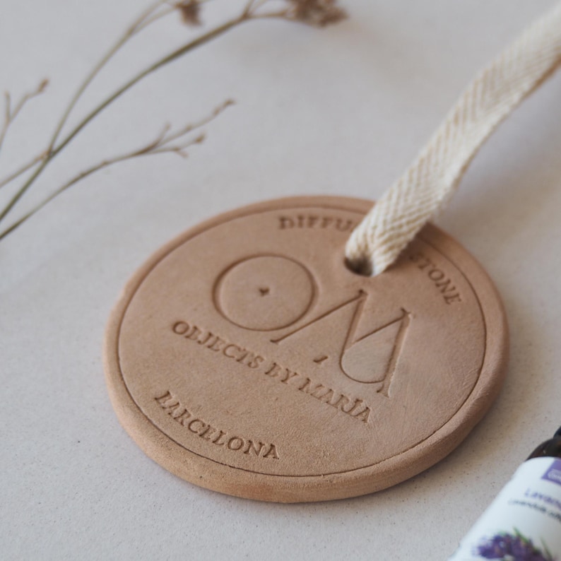 May include: A circular, light brown diffuser stone with the embossed text "DIFFUSER STONE", "OM", "OBJECTS BY MARIA", and "BARCELONA". It has a cream-coloured ribbon for hanging. The image is of an aromatherapy product.