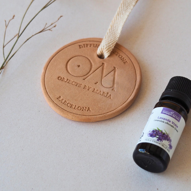 May include: A circular, tan diffuser stone with the text "DIFFUS STONE" and "OBJECTS BY MARIA BARCELONA". A small bottle of lavender essential oil is next to the diffuser stone. A beige ribbon is attached to the diffuser stone.