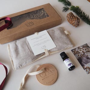 May include: A gift set including a brown cardboard box, a lavender-scented eye pillow, a small bottle of lavender essential oil, and a leather tag. The eye pillow is labelled "ALMOHADILLA RELAJANTE PARA OJOS". The set includes a pine cone and sprig.