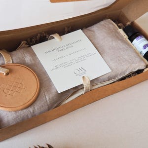 Può includere: Una scatola regalo rettangolare in cartone contenente un cuscino per occhi in lino beige, un disco rotondo in terracotta con motivo a griglia e una piccola bottiglia di vetro di olio essenziale di lavanda. Una carta bianca con testo in spagnolo.