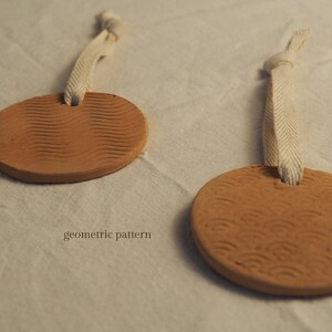 May include: Two terracotta-coloured, circular ornaments with textured geometric patterns. Each ornament has a cream-coloured fabric loop for hanging. The text "geometric pattern" is visible on the surface.