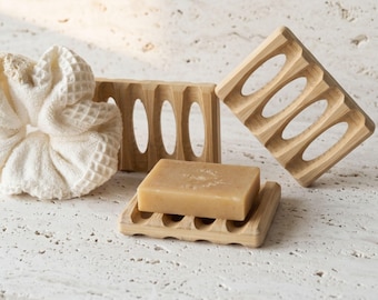 May include: Three light-colored wooden soap dishes with oval cutouts, one holding a rectangular beige bar of soap, accompanied by a white waffle-textured fabric scrunchie. The wooden dishes feature a slatted design for drainage.