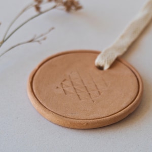 Puede incluir: Adorno redondo de cerámica color terracota con un diseño geométrico texturizado. Se adjunta un lazo de tela color crema para colgarlo. El adorno mide aproximadamente 7,6 cm de diámetro.