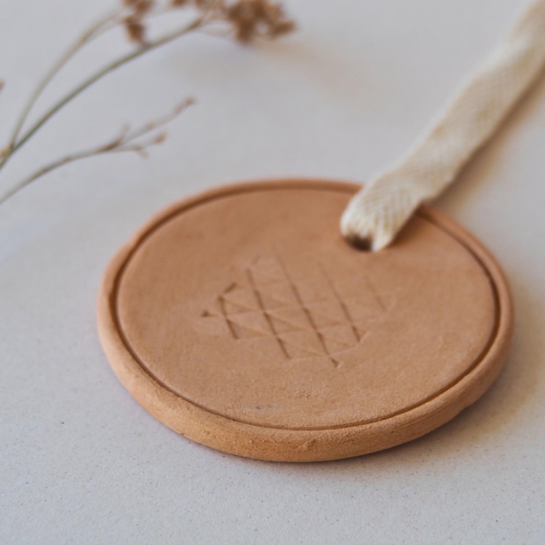 May include: A circular, terracotta-coloured ceramic ornament with a textured geometric design. A cream-coloured fabric loop is attached for hanging. The ornament is approximately 7.6 cm in diameter.