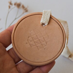 May include: A circular, terracotta-coloured ornament with a geometric pattern and a cream ribbon for hanging. The ornament has a textured surface and a minimalist design. The pattern is made up of small, interconnected triangles.