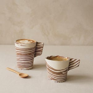 Pode incluir: Duas canecas de cerâmica com um padrão de riscas horizontais castanhas e creme marmoreadas e uma pega angular distinta. A caneca mais alta contém um latte com arte em espuma, enquanto a mais curta contém café preto. Uma colher de madeira.