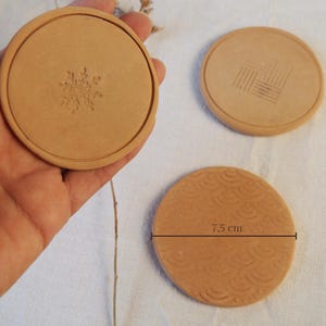 May include: Three round, light brown ceramic coasters. One has a snowflake design, another a geometric pattern, and the third a wave pattern. The coaster with the wave pattern is 7.5 cm in diameter.