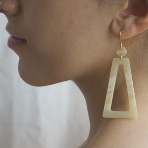 Peut inclure: Une paire de boucles d'oreilles géométriques de couleur crème avec un crochet doré. Les boucles d'oreilles sont faites d'un matériau qui ressemble à de l'os ou de la coquille et ont un motif rayé.