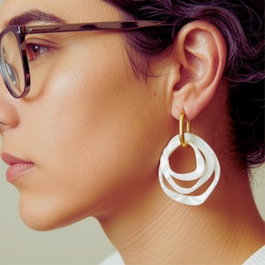 May include: A gold-toned metal hoop earring featuring three interconnected, wavy white circular charms. The earring is suspended from a small, polished gold-colored oval link that attaches directly to the earlobe, creating a layered, organic aesthetic.