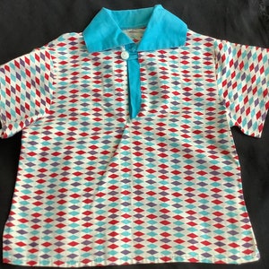 Può includere: Una camicia a maniche corte con un motivo a rombi rosso, blu e viola su sfondo bianco. La camicia ha un colletto blu e una chiusura con bottone.