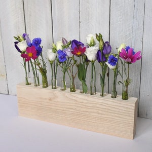 May include: A wooden flower vase with a light wood finish holds a variety of flowers in different colors, including white, pink, blue, and purple. The flowers are arranged in a row in the vase, with the stems extending out of the top.