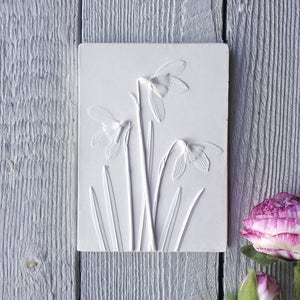 May include: A white rectangular relief sculpture featuring a detailed depiction of snowdrop flowers. The artwork showcases the delicate petals and stems of the flowers, creating a textured, three-dimensional effect. The sculpture is set against a light gray wooden background.