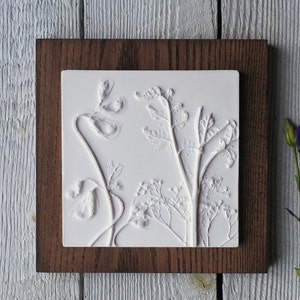 May include: A white plaster wall art piece with a pressed floral design. The design features delicate flowers and leaves in a natural, organic style. The piece is framed in a dark wood frame.