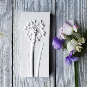 May include: White plaster wall art featuring a raised relief of a flower with a round head and long stem. The flower is similar to an allium or onion blossom. The wall art is next to a bouquet of purple and white sweet pea flowers.