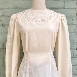 May include: Cream-coloured blouse with long sleeves and a delicate lace collar. The blouse has a fitted waist and a subtle sheen, suggesting a satin or silk fabric. The sleeves are gathered at the cuffs, adding a touch of vintage style. The blouse is paired with blue jeans.