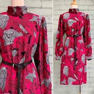 May include: A vintage dress in fuchsia, with long sleeves and a high neck. It features a black belt and a paisley and floral pattern in black and white. The dress is displayed on a mannequin. The dress's length is approximately 100 cm.