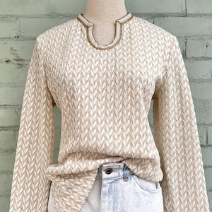 May include: Cream-colored long-sleeve sweater with a textured, chevron-like pattern. The neckline is trimmed with white and gold detailing. The sweater is paired with light-wash denim jeans.