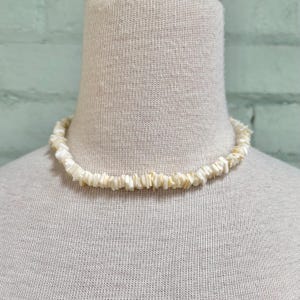 May include: An off-white shell chip necklace. The necklace is made of small, rectangular shell pieces strung together, creating a textured, natural appearance. The necklace is displayed on a mannequin.