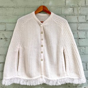 Op de afbeelding: Een witte gebreide cape met knoopsluiting en een franje rand. De cape is gemaakt van een lichtgewicht, opengeweven stof.