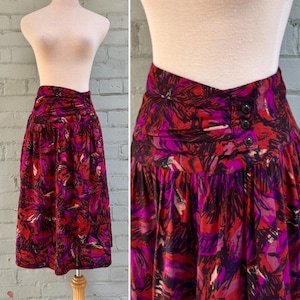 May include: A vintage midi skirt with a vibrant floral pattern in shades of red, purple, and black. The skirt features a high waist with three decorative buttons and a pleated design, displayed on a mannequin.