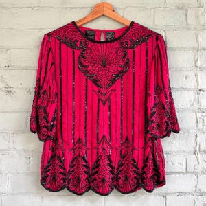 May include: A vibrant pink vintage top adorned with black sequins and beads. It has a round neckline, three-quarter sleeves, and a scalloped hem. The design incorporates vertical stripes and decorative patterns.
