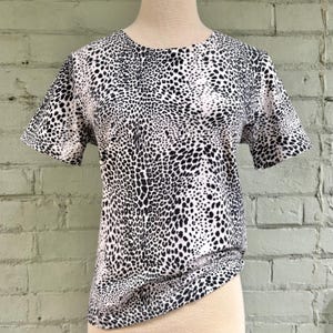May include: A short-sleeved, crew neck t-shirt with a black and white animal print pattern. The shirt is displayed on a mannequin and is made of a soft, lightweight fabric. The pattern is a repeating design of black spots on a white background.