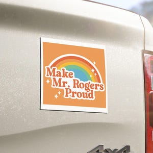 May include: A square magnet with an orange background and a rainbow design. The text "Make Mr. Rogers Proud" is displayed in a retro font. The magnet is attached to the back of a vehicle.