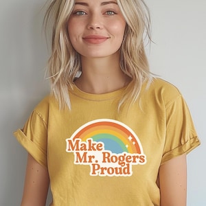 May include: Mustard yellow t-shirt with rolled-up sleeves. The shirt features a rainbow graphic and the text "Make Mr. Rogers Proud" in a retro font. The shirt is made of a soft material.