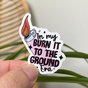 May include: A pink and white sticker with a cartoon lighter and the text "In my burn it to the ground Era".