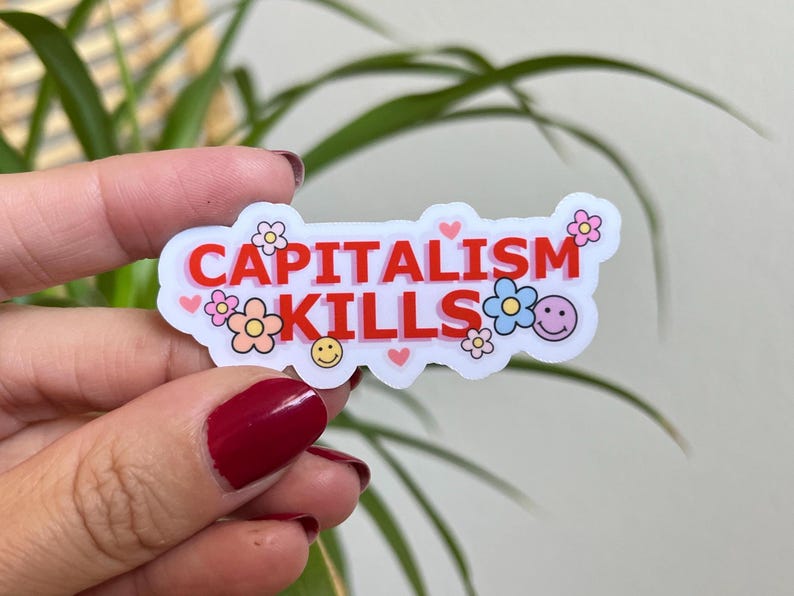 May include: A white sticker with the words "CAPITALISM KILLS" in red, surrounded by flower and heart graphics. The sticker is held up in front of a green plant.