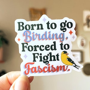 A die-cut sticker featuring the text "Born to go Birding, Forced to Fight Fascism." The words are printed in black. A small yellow and black goldfinch illustration is on the right.
