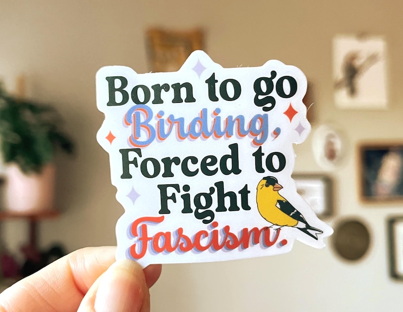 A die-cut sticker featuring the text "Born to go Birding, Forced to Fight Fascism." The words are printed in black. A small yellow and black goldfinch illustration is on the right.