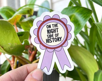 May include: A sticker with a light purple ribbon design and the text "ON THE RIGHT SIDE OF HISTORY" in black. The sticker has a white background and a scalloped edge. The sticker is held in front of green leaves.