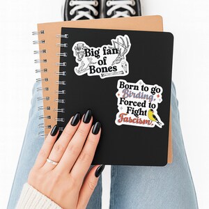 Puede incluir: Un cuaderno de espiral negro con dos pegatinas. La pegatina superior muestra dibujos lineales blancos de cr&aacute;neos y huesos de animales con el texto "Big fan of Bones". La pegatina inferior muestra un p&aacute;jaro amarillo y el texto "Born to go Birding, Forced to Fight Fascism".