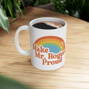 May include: White ceramic coffee mug with a curved handle, filled with dark coffee. The mug features a retro rainbow design and the text "Make Mr. Rogers Proud" in orange lettering. The mug sits on a wooden surface.